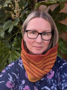 Dalton Cowl in Autumnal colours being worn by a blonde lady wearing glasses who is stood in front of a leafy plant and she is wearing a blue floral dress.