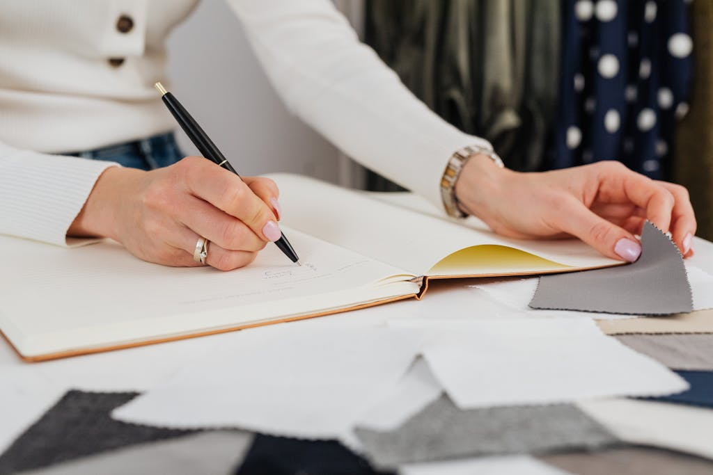Close-up of hands sketching in a notebook, selecting fabric samples for fashion design.