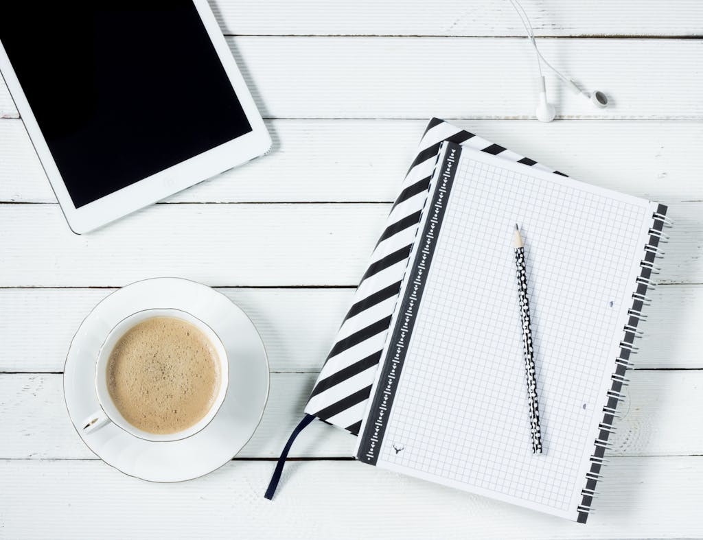 Flatlay of a modern workspace featuring coffee, tablet, and notebook with pencil on a white wooden desk.